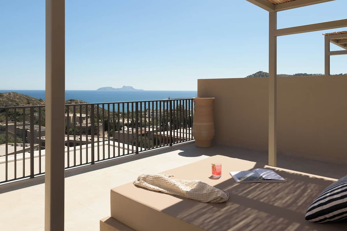 A balcony with a view of the sea, a table with a drink and a book, and a woven ceiling.
