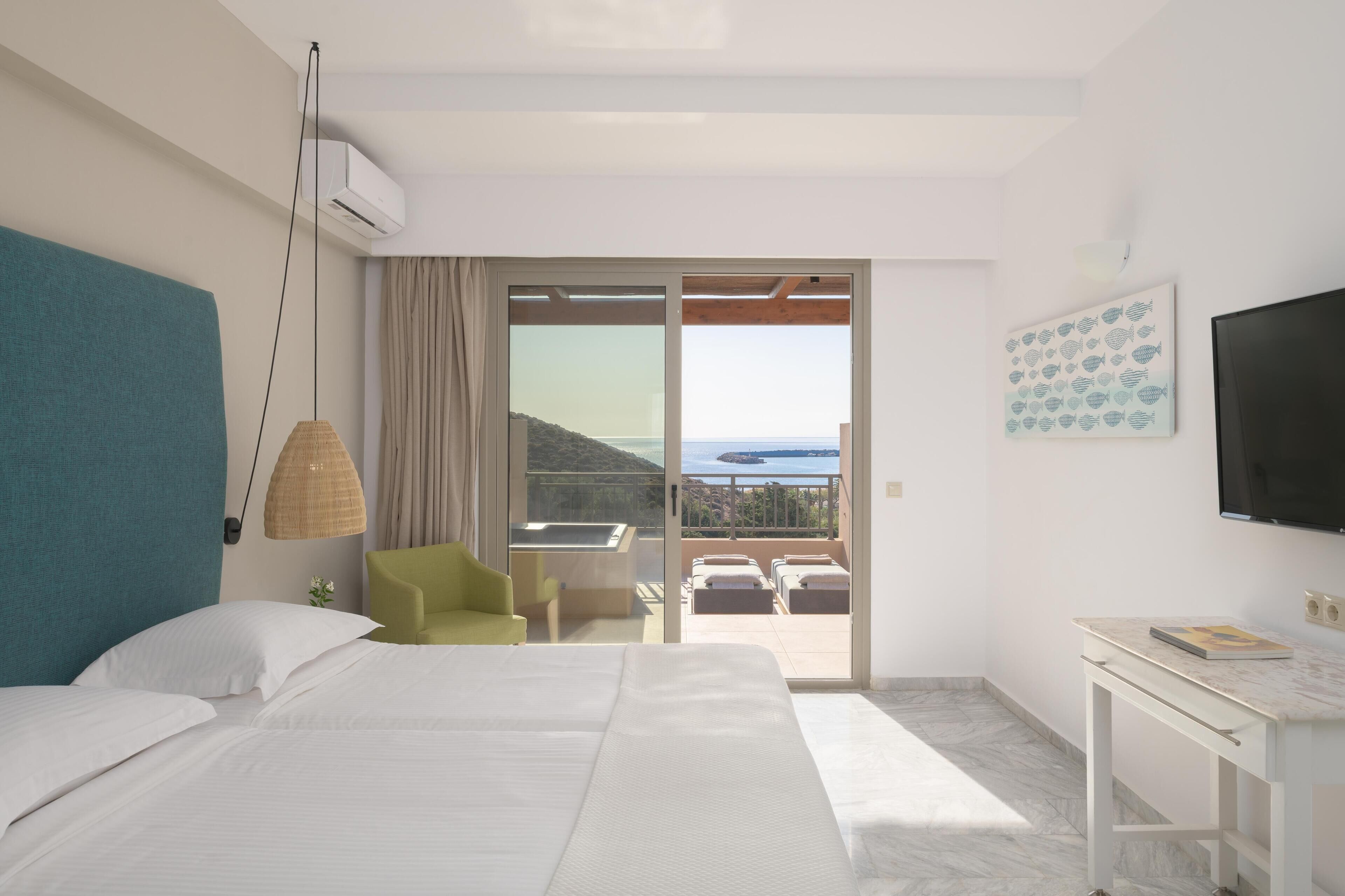 Habitación de hotel con cama, escritorio, silla, televisor y balcón con vista al mar.