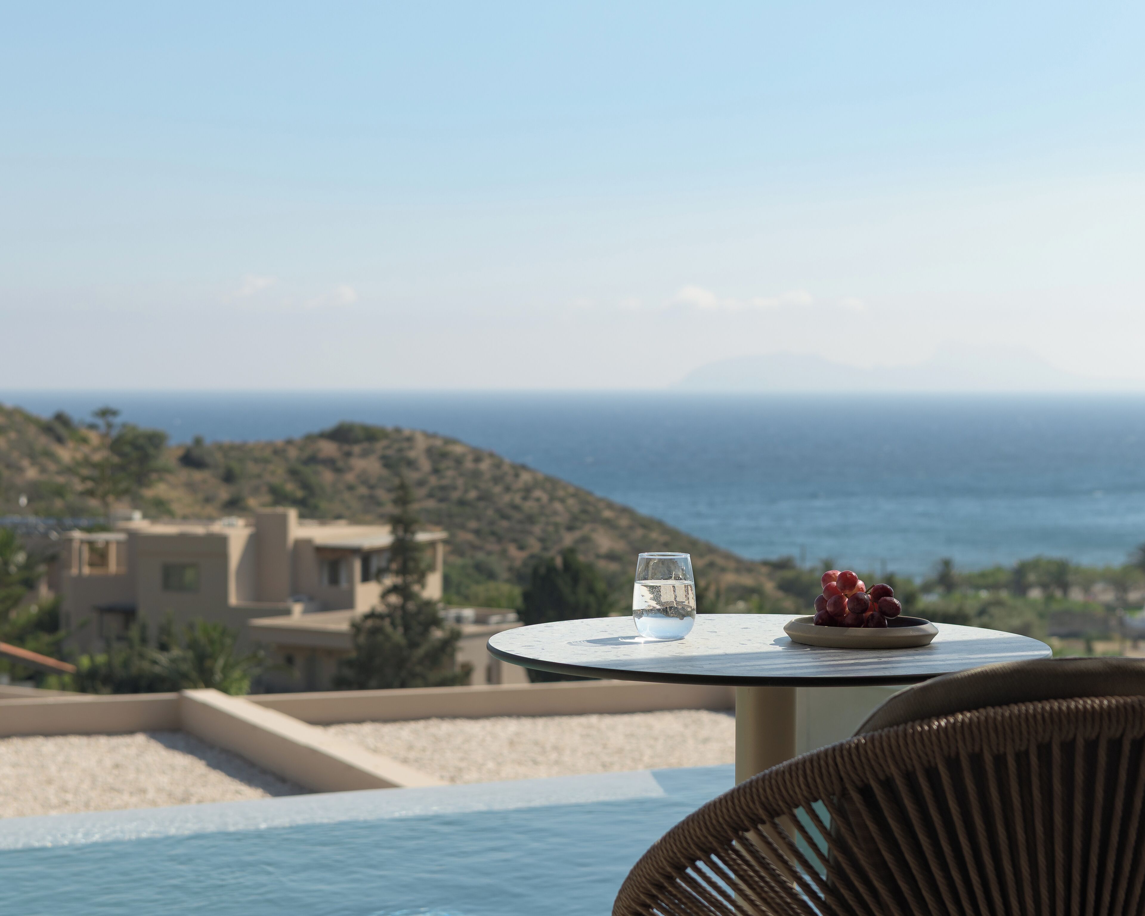 Una mesa con un vaso de agua y uvas, con vista a una piscina y al mar y las colinas.