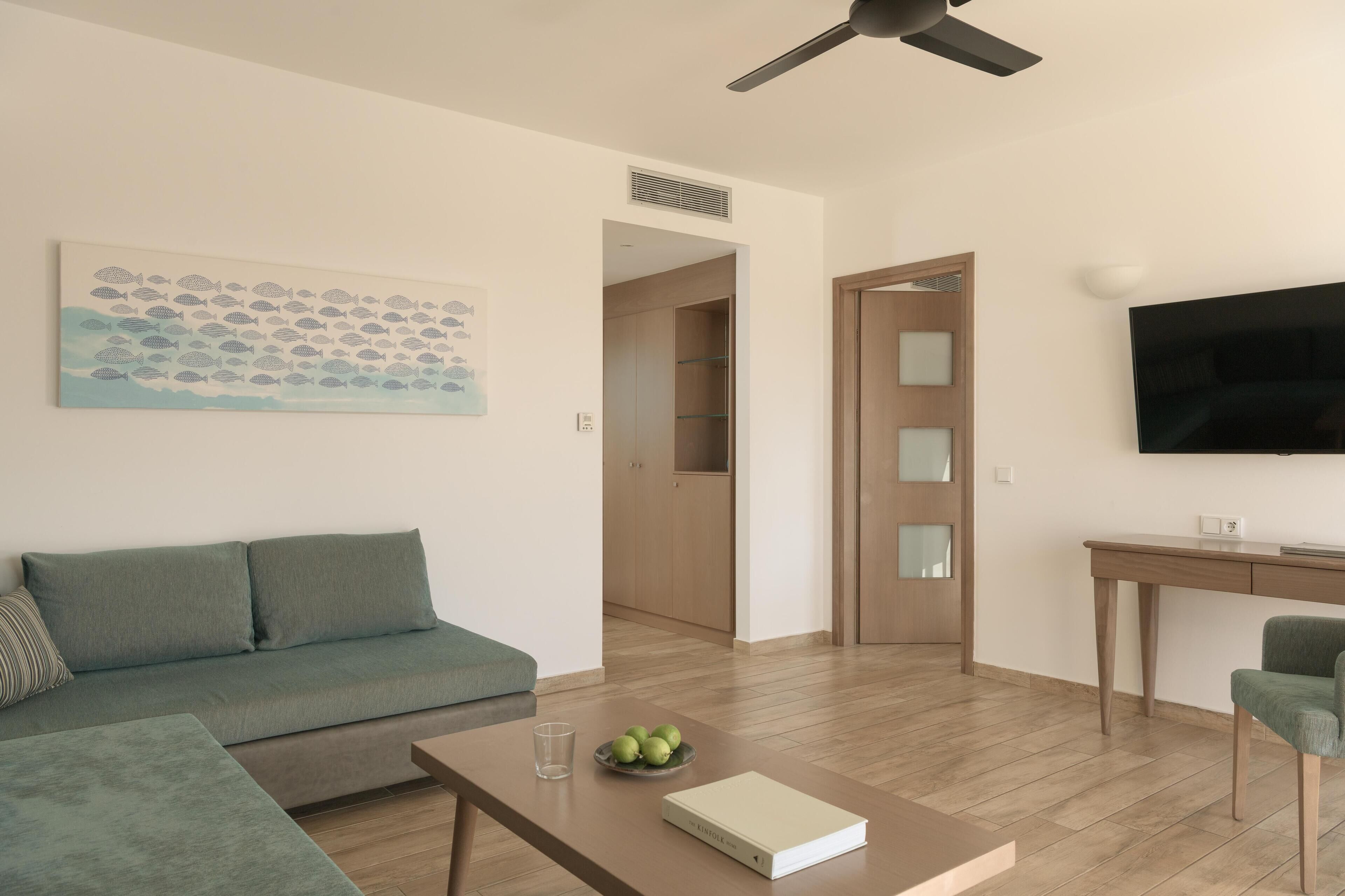 Una sala moderna con un sofá, una mesita de centro con un libro y fruta, un televisor y un cuadro en la pared.