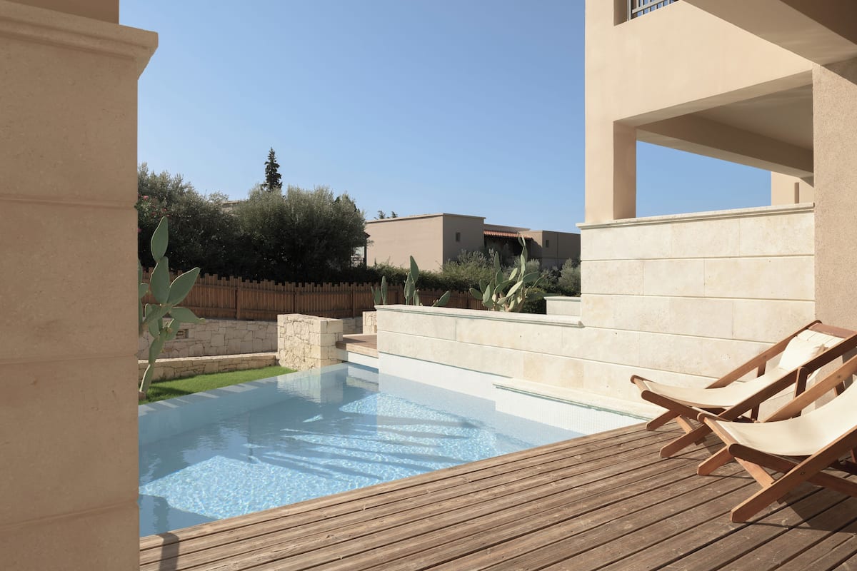 A swimming pool with a wooden deck and lounge chairs, surrounded by a low stone wall and greenery.