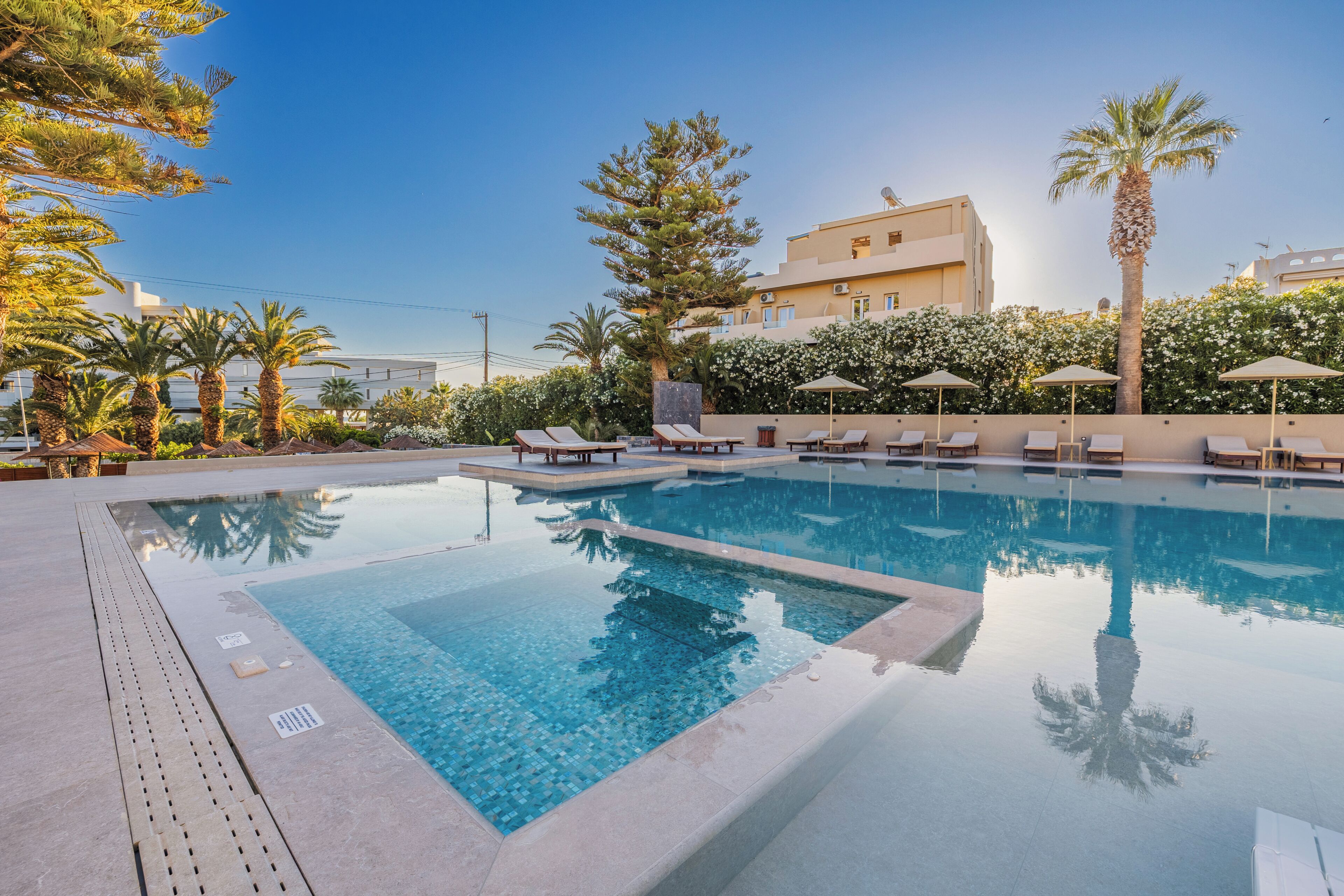 A swimming pool with lounge chairs and umbrellas, surrounded by palm trees and a building with a beige facade.
