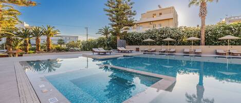 A swimming pool with lounge chairs and umbrellas, surrounded by palm trees and a building with a beige facade.