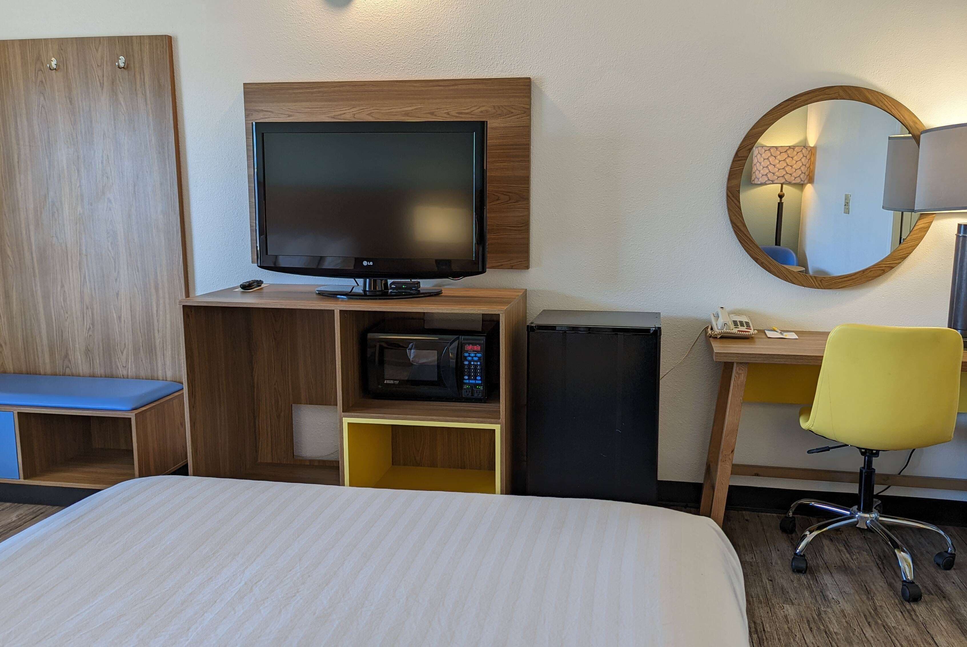 A hotel room with a bed, a TV, a microwave, a desk with a chair, and a mirror.