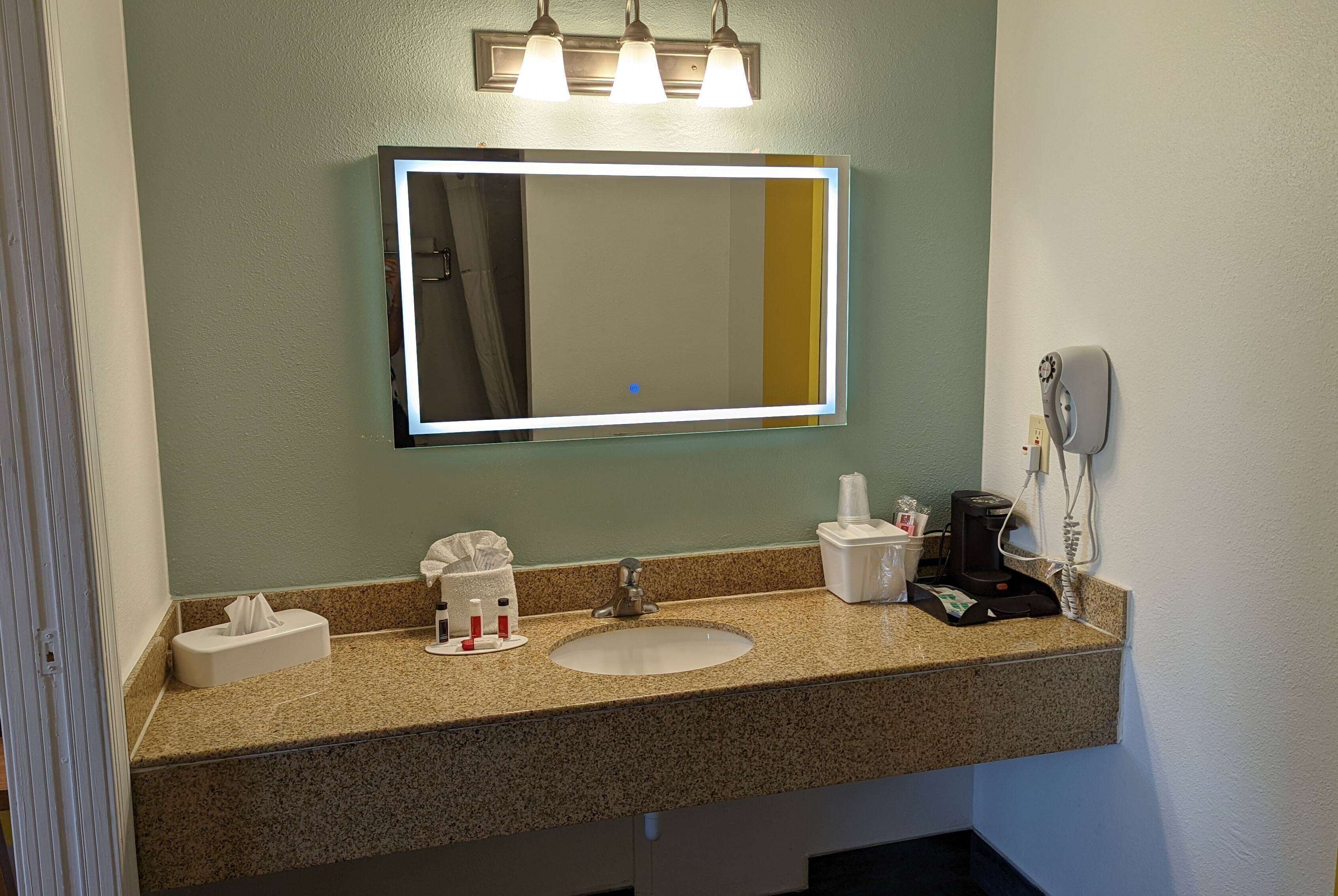 A bathroom with a granite countertop, a white sink, a mirror with lights, a tissue box, a soap dispenser, a toothbrush holder, a cup, a coffee maker, and a phone.