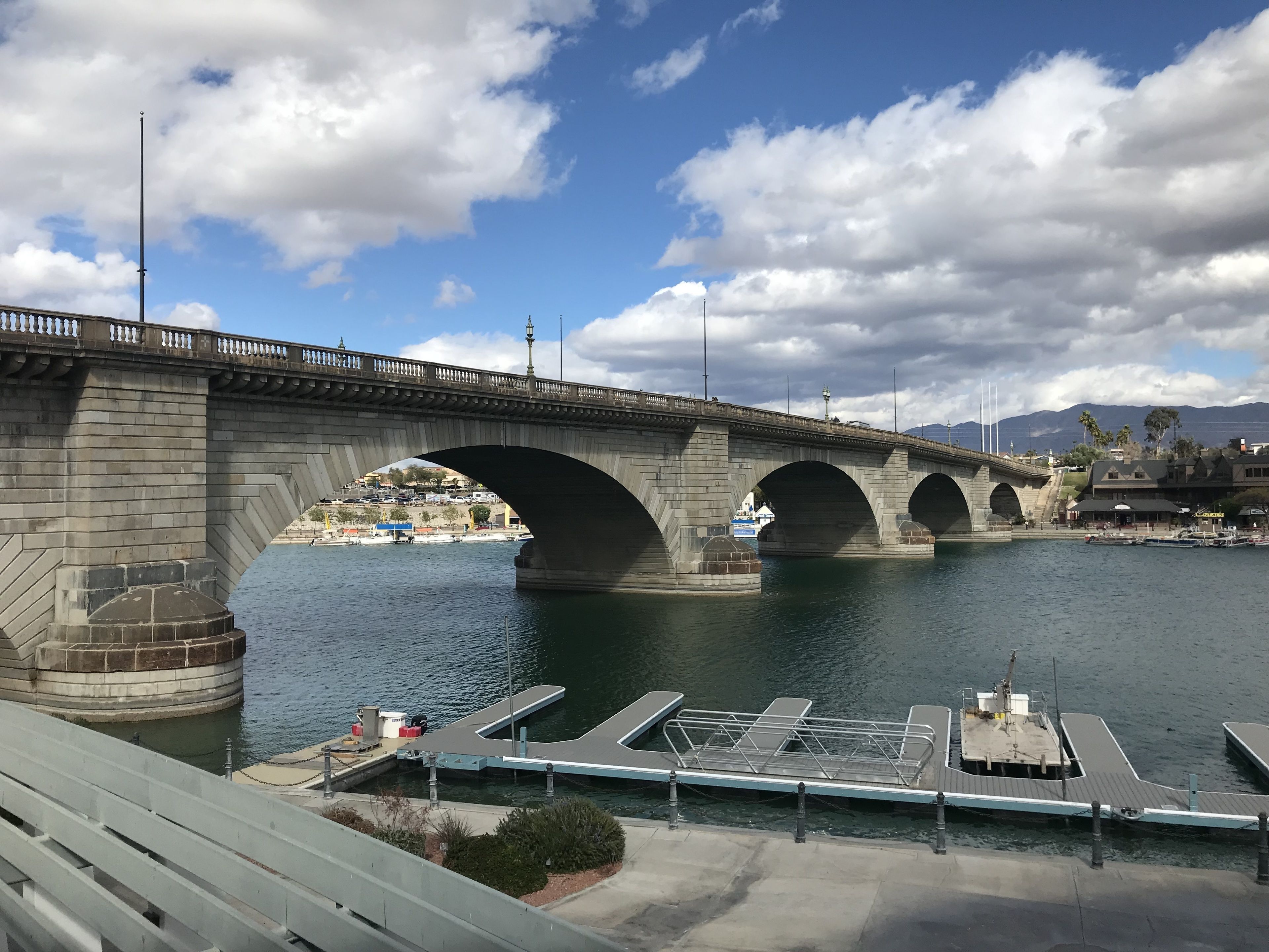 A stone bridge with multiple arches over a body of water, leading to a dock with boats and a marina.