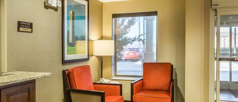 A hotel lobby with two orange chairs, a desk with a telephone, a framed picture on the wall, and a window with a view of the street.