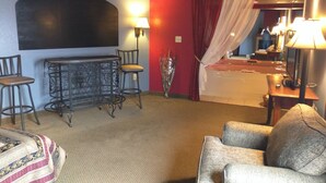 A hotel room with a bed, a sitting area with a chair and a small table, a bar area with stools, and a bathroom with a bathtub and a mirror.