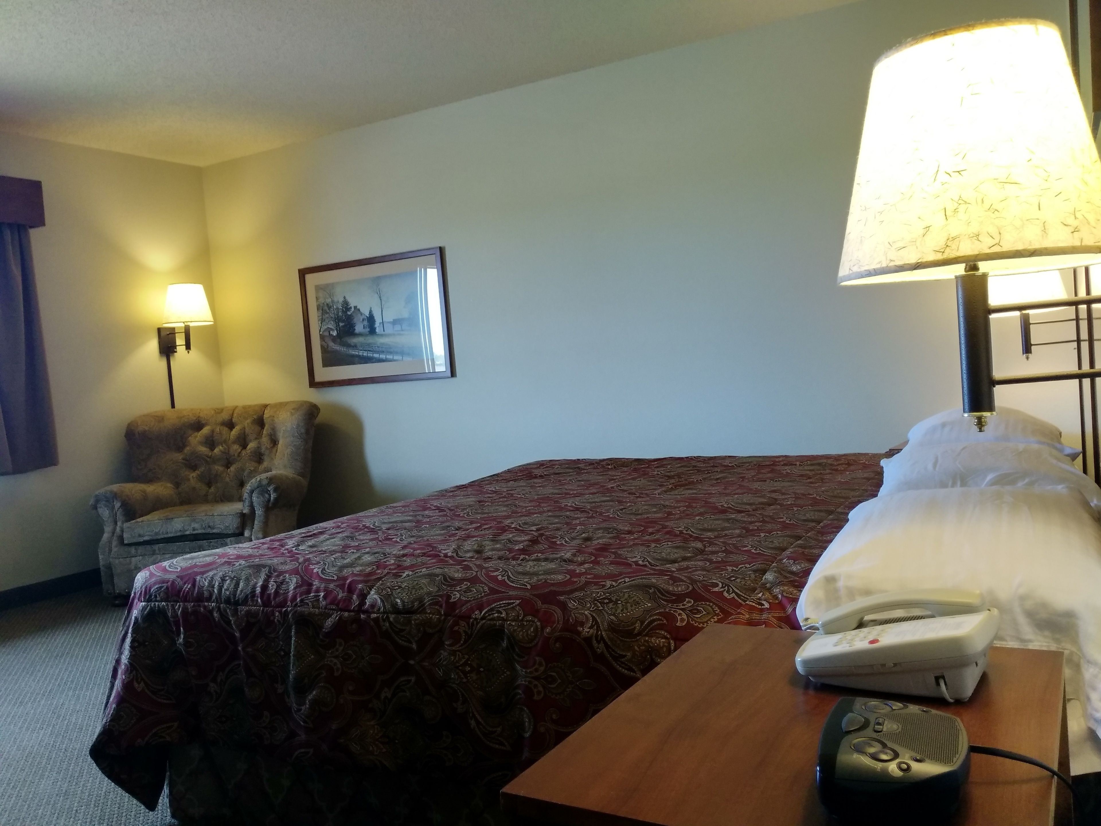 Quarto de hotel com cama, poltrona, mesa, abajur e telefone.