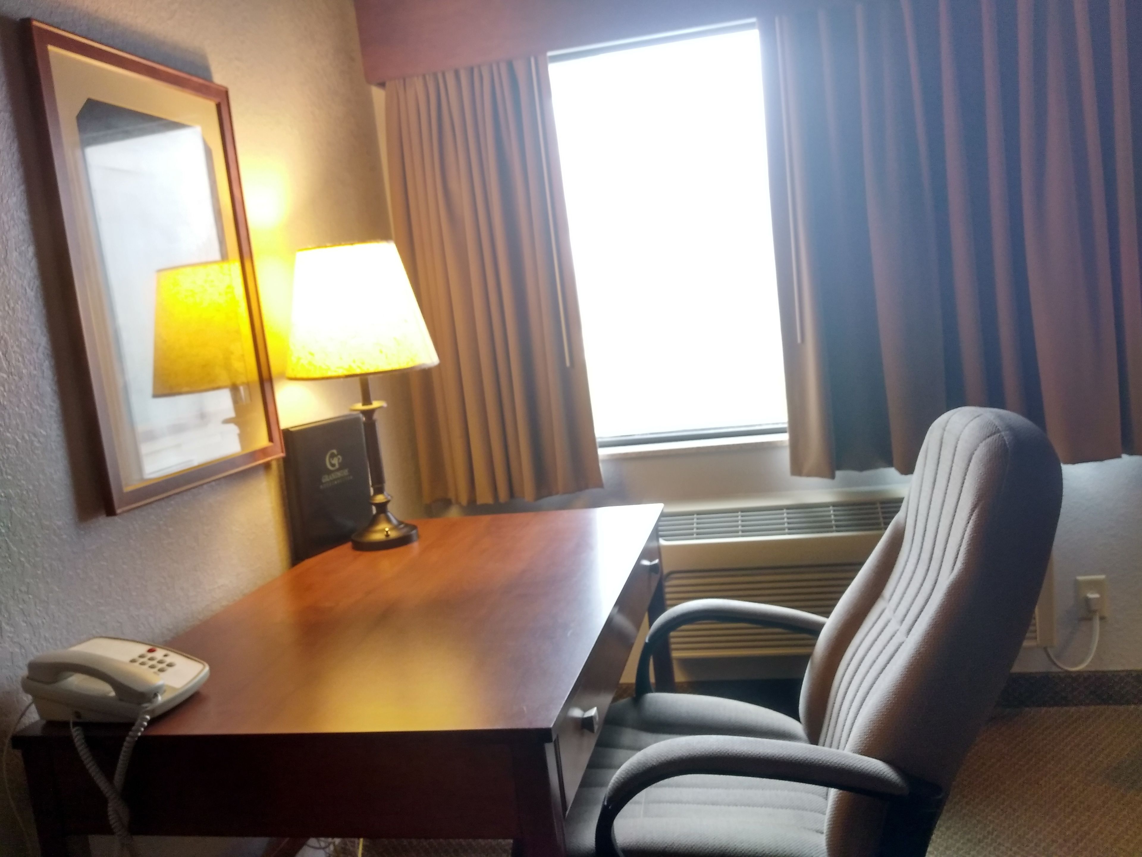 Habitación de hotel con un escritorio de madera, una lámpara, una silla, un teléfono y una ventana con cortinas.