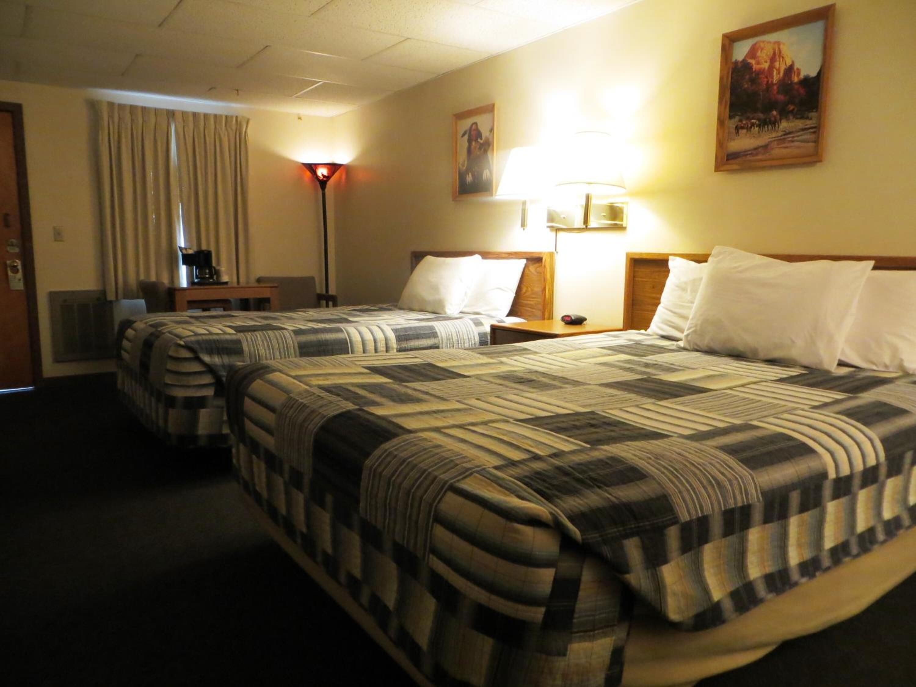 A hotel room with two beds, a desk, a chair, a lamp, and a painting on the wall.