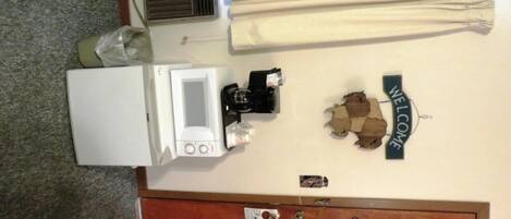 A room with a white refrigerator, a coffee maker, a 'WELCOME' sign, a wooden door, and a window with blinds.