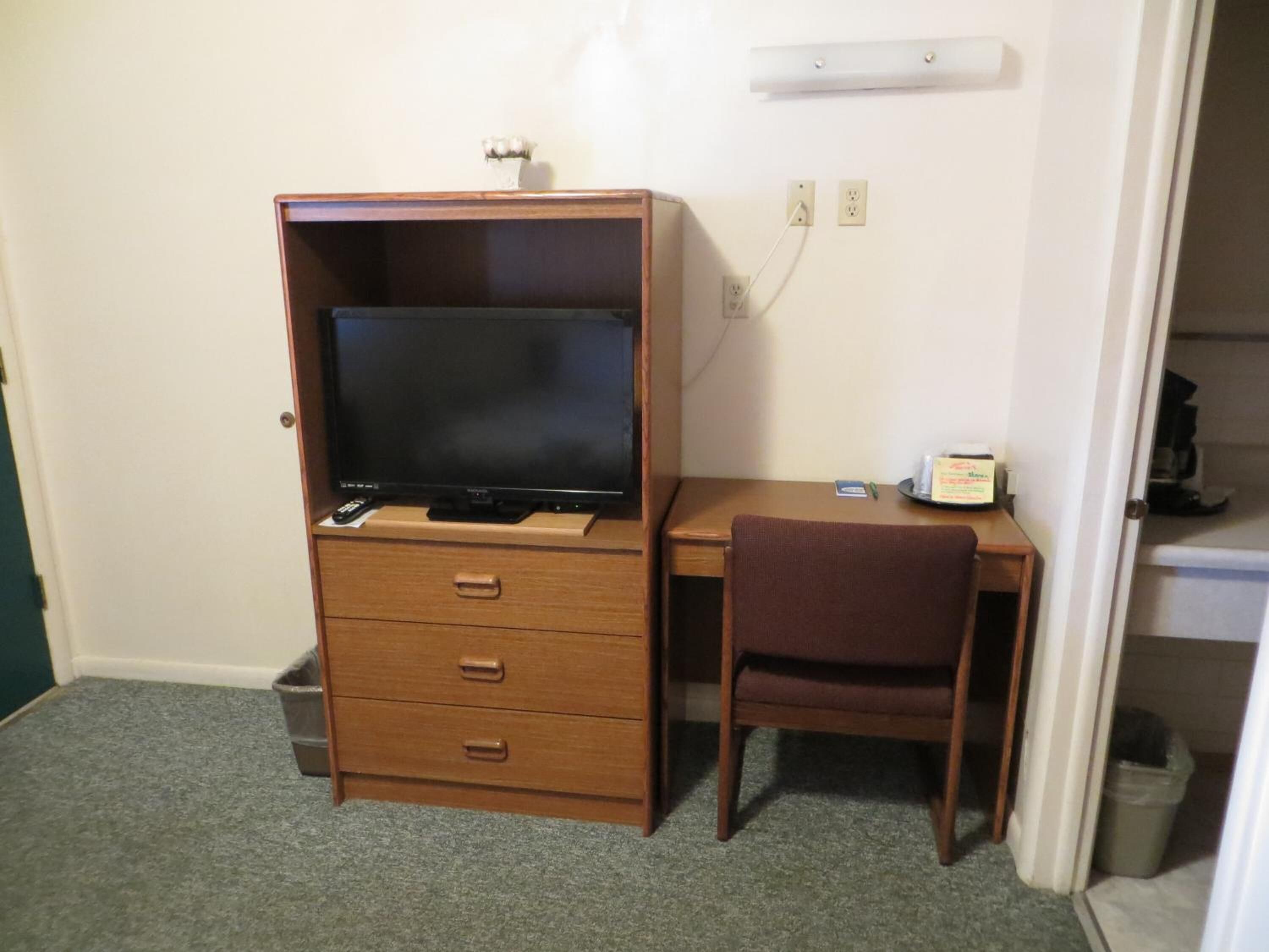 Una habitación pequeña con un escritorio de madera, un televisor sobre un soporte, una silla y un cubo de basura.