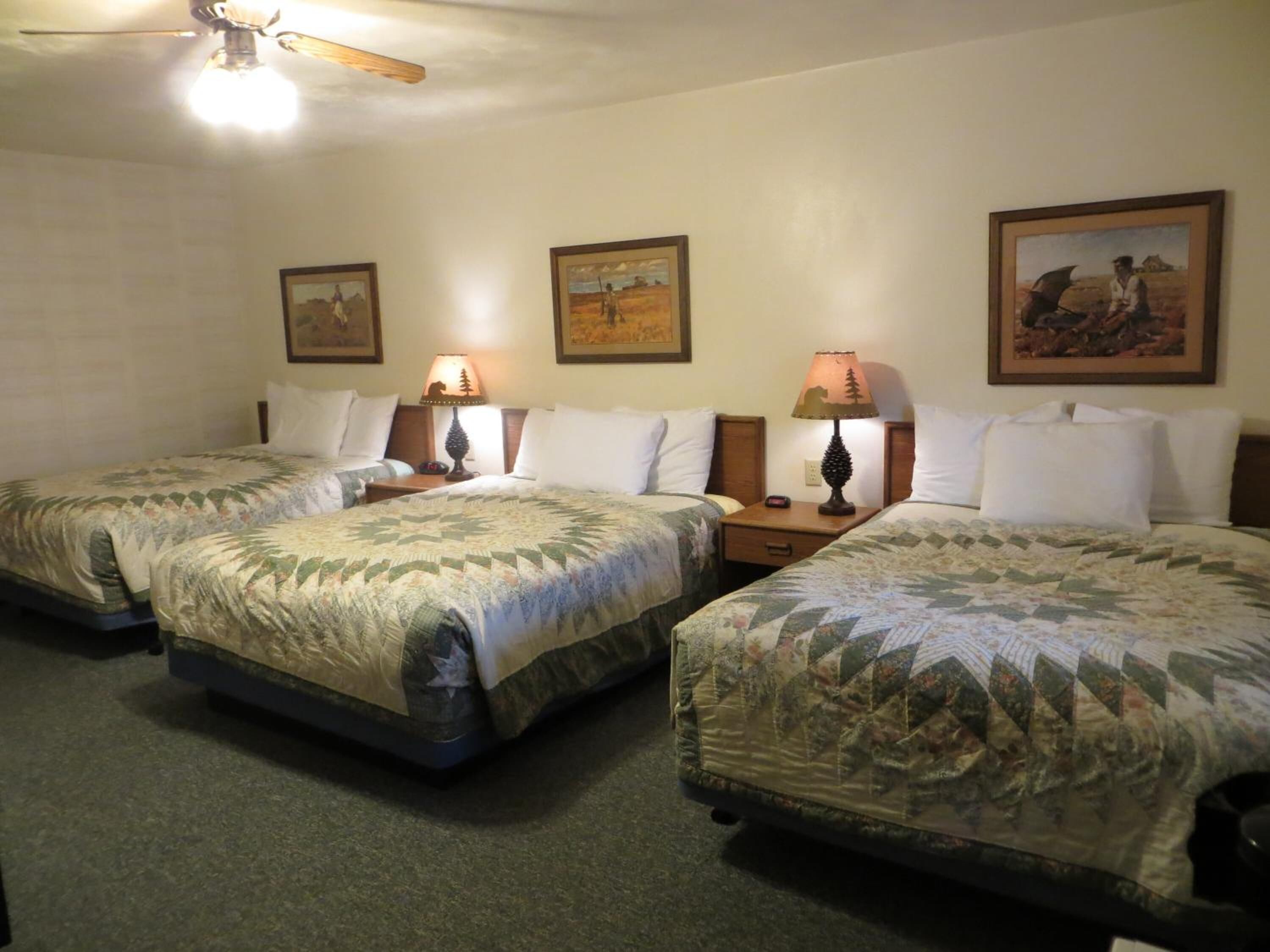 A hotel room with three single beds, each with a quilted bedspread. There are two wall-mounted lamps on wooden nightstands, and a ceiling fan above.