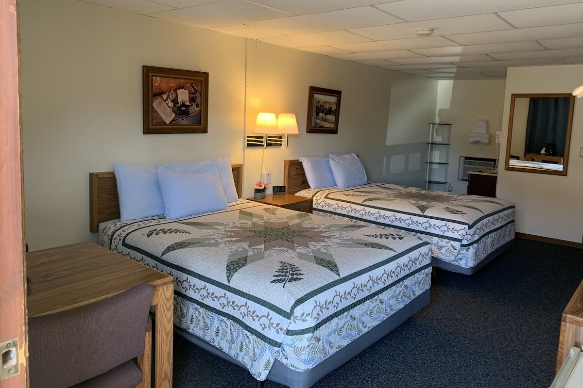 A hotel room with two beds, a desk, a chair, a lamp, a mirror, and a shelf with towels.