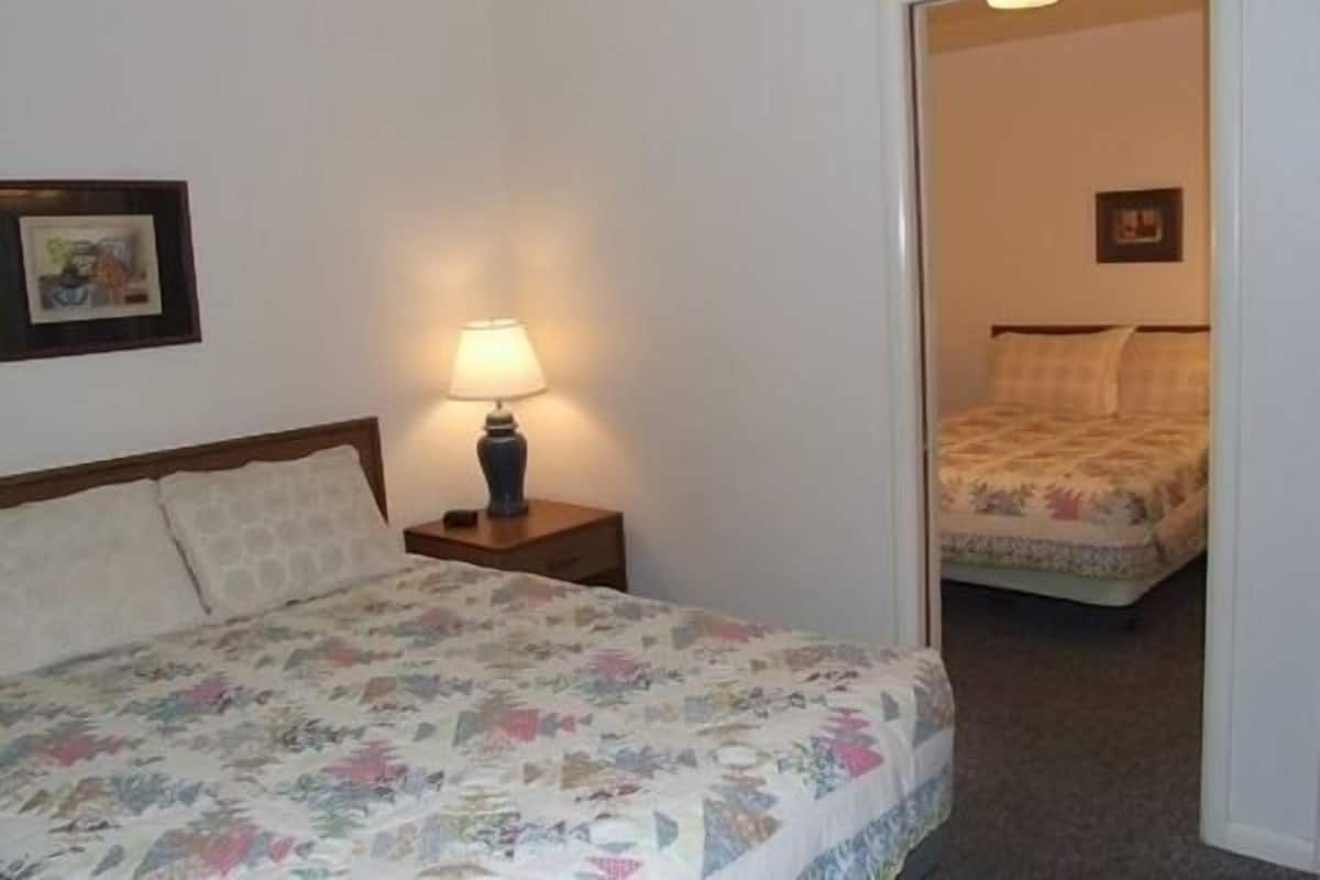A hotel room with a bed, a nightstand with a lamp, and a door leading to another room.