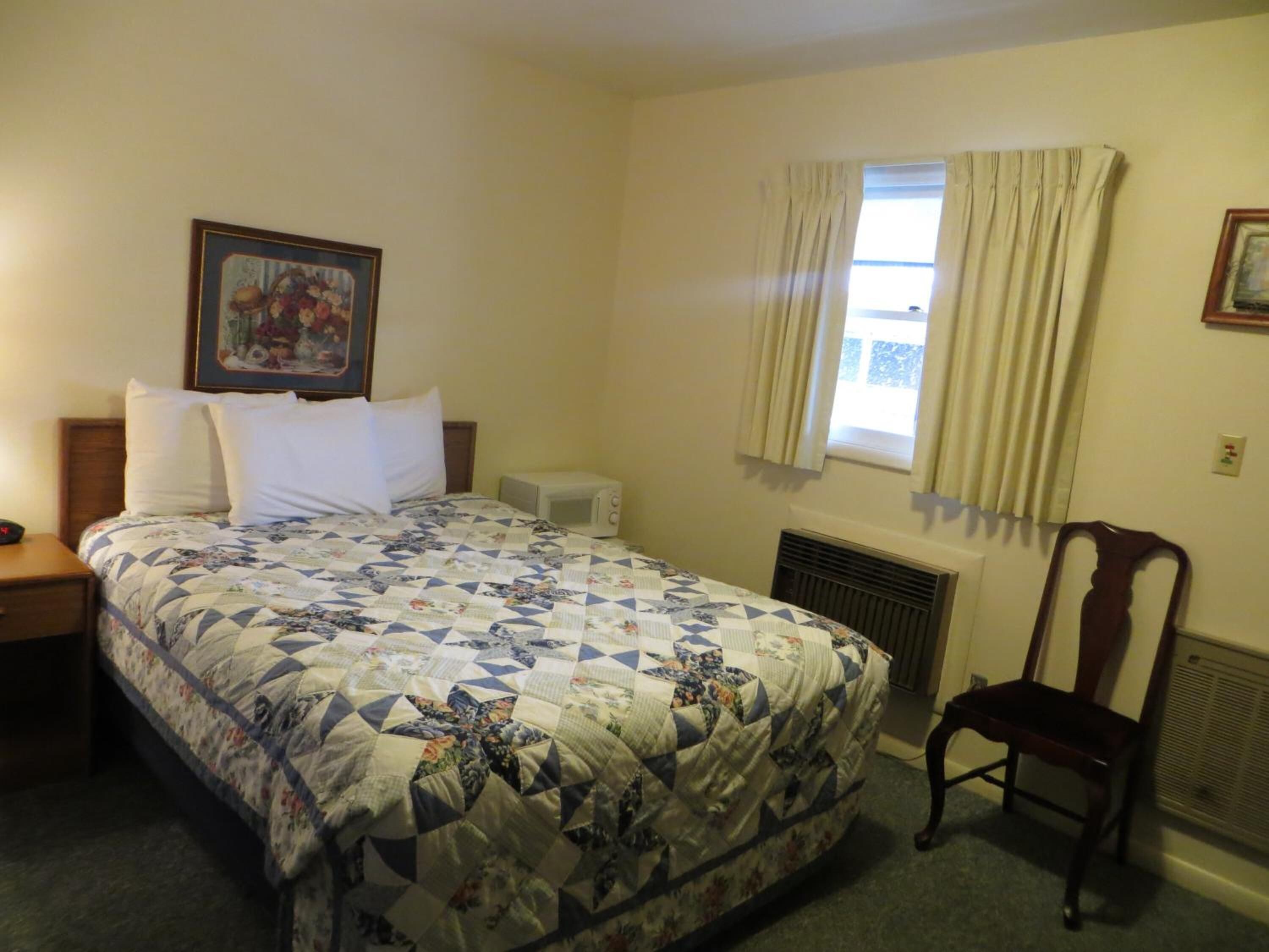 A hotel room with a bed, a chair, a picture on the wall, a window with curtains, and a radiator.
