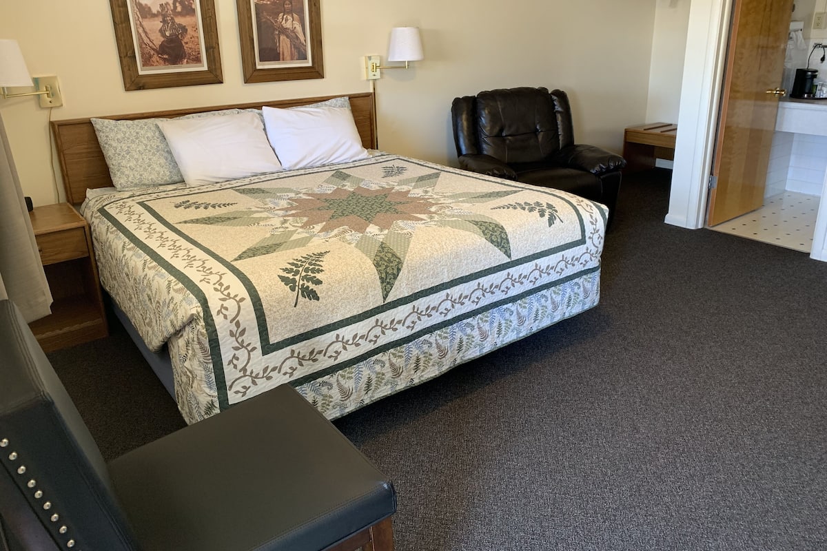 A neatly made bed with a quilt, a chair, and a small table in a room with a carpeted floor.