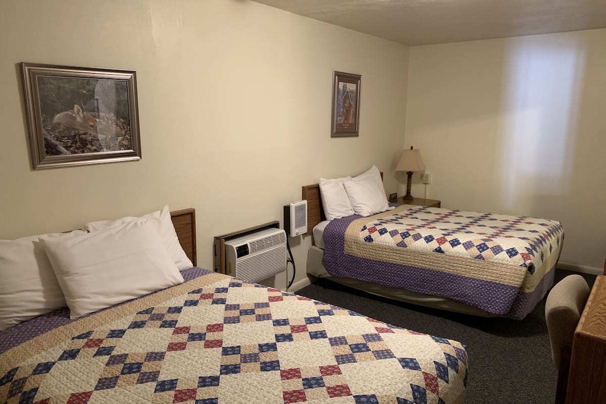 A hotel room with two beds, each with a quilted bedspread, a wall-mounted air conditioner, and two framed pictures on the wall.