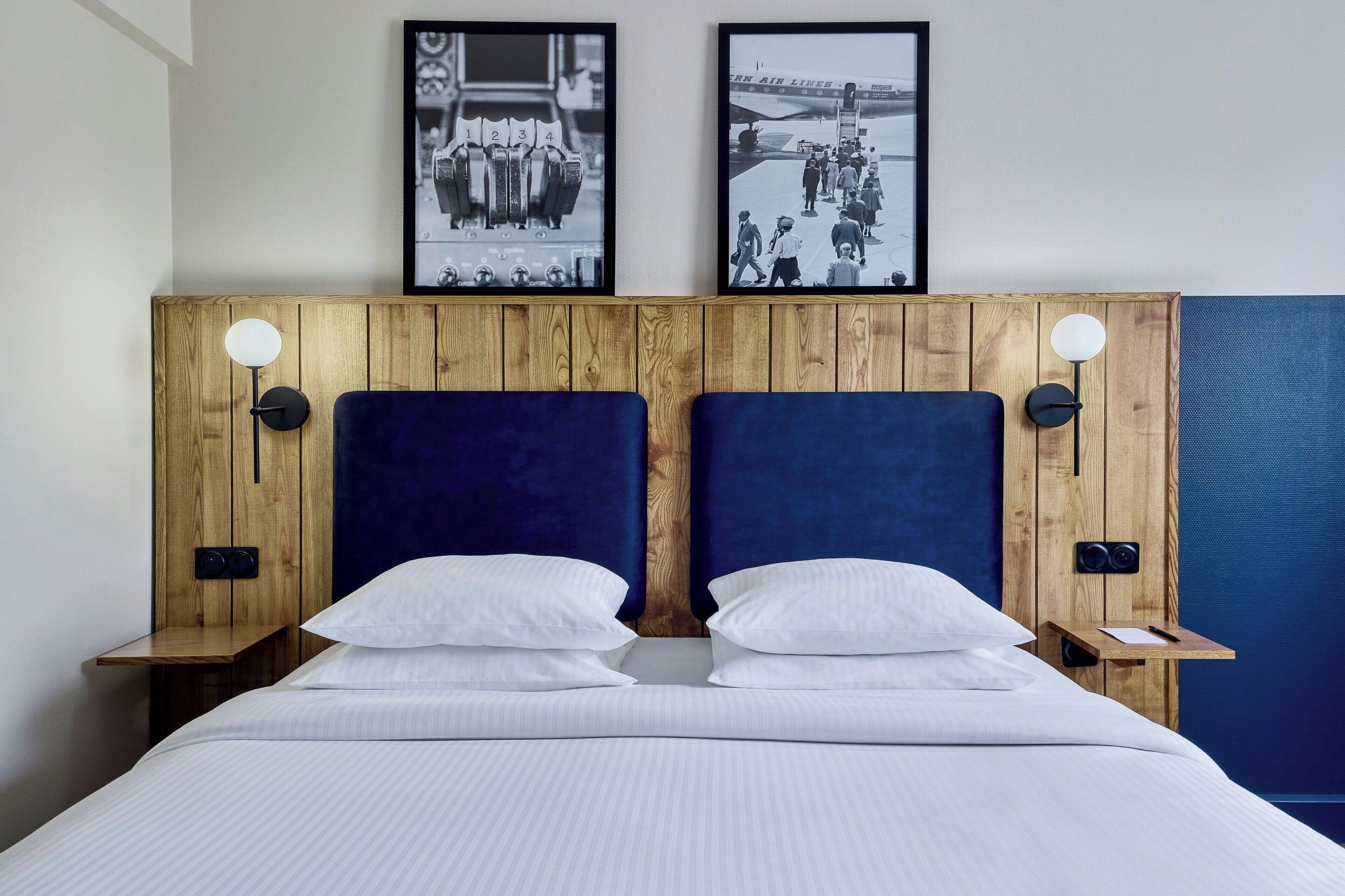 A double bed with white linens, a wooden headboard with two wall-mounted lights, and two framed black-and-white photographs above the bed.