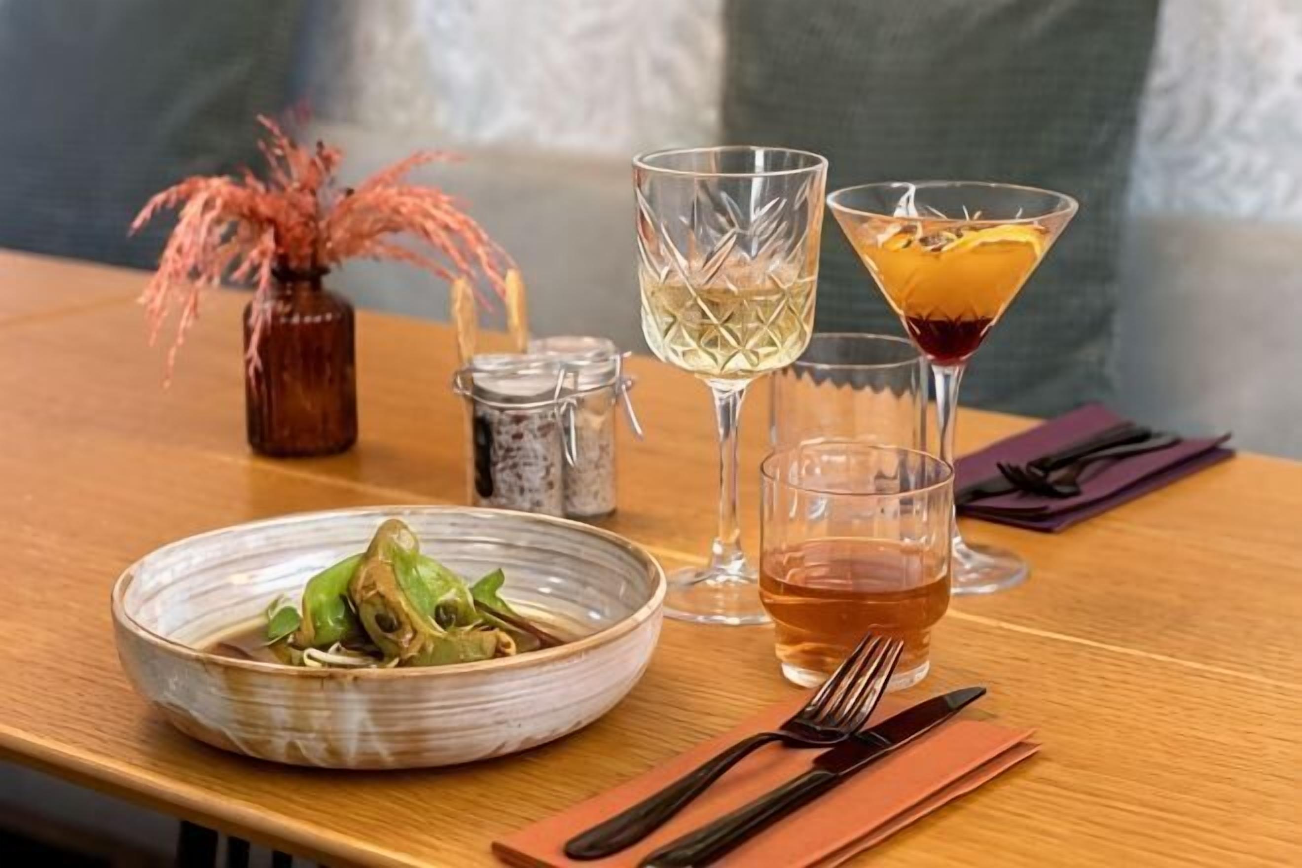 A dining table with a bowl of food, two glasses of a clear beverage, a martini glass with a dark drink, and a small vase with dried flowers.