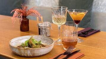A dining table with a bowl of food, two glasses of a clear beverage, a martini glass with a dark drink, and a small vase with dried flowers.