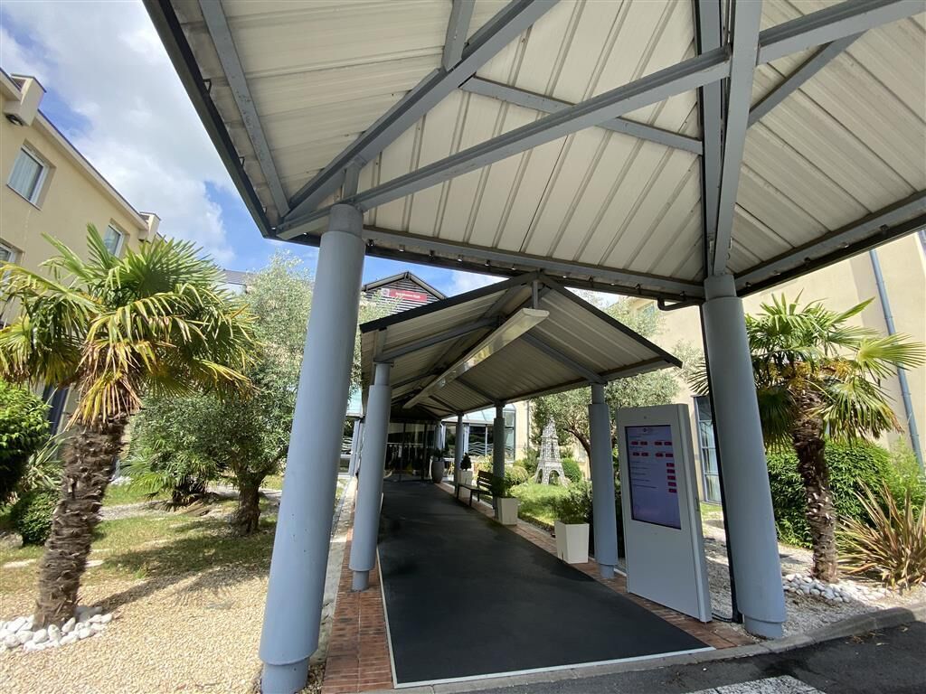 A covered walkway with palm trees and a bench, leading to a building entrance.