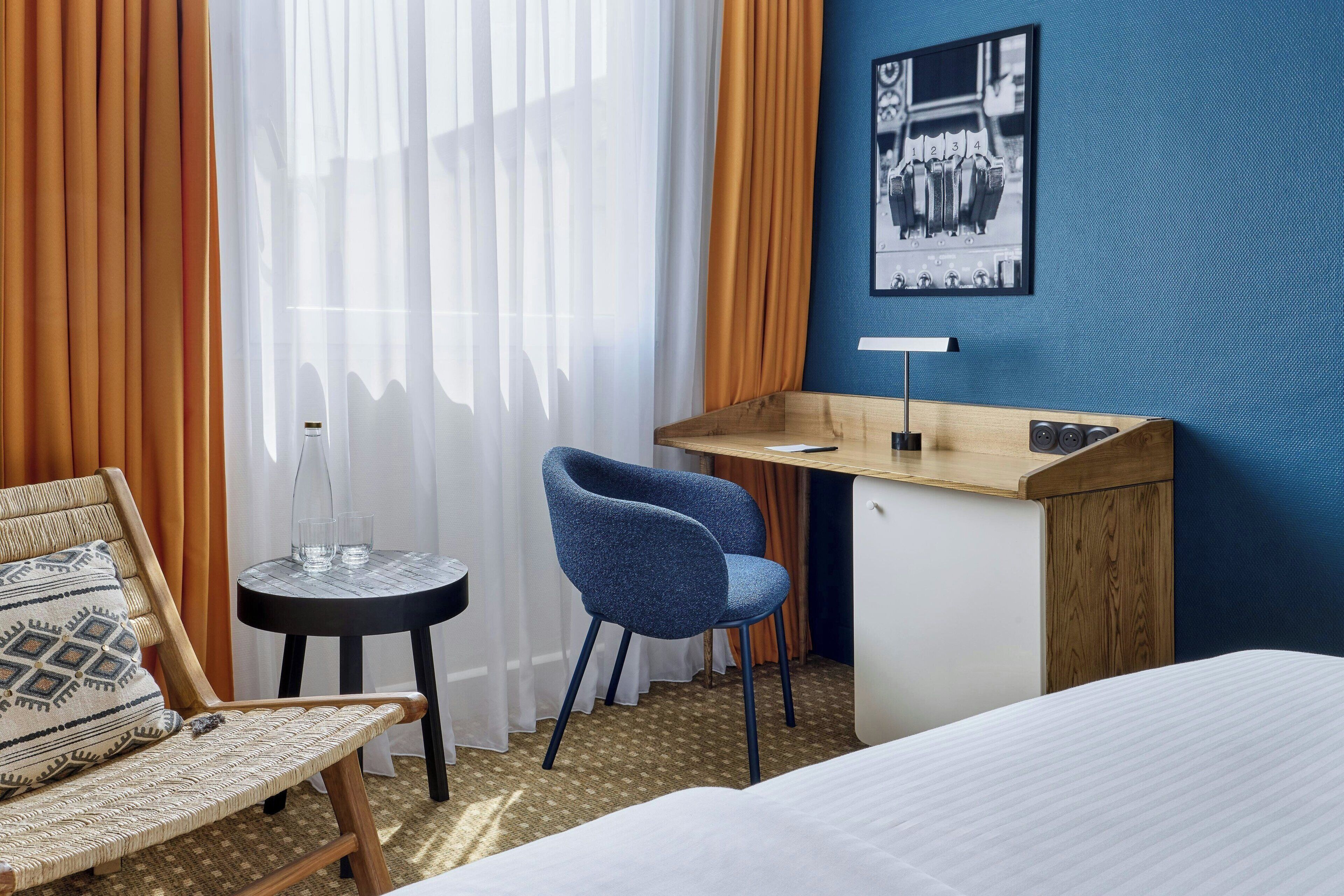 A hotel room with a bed, a desk, a chair, a small table, and curtains.