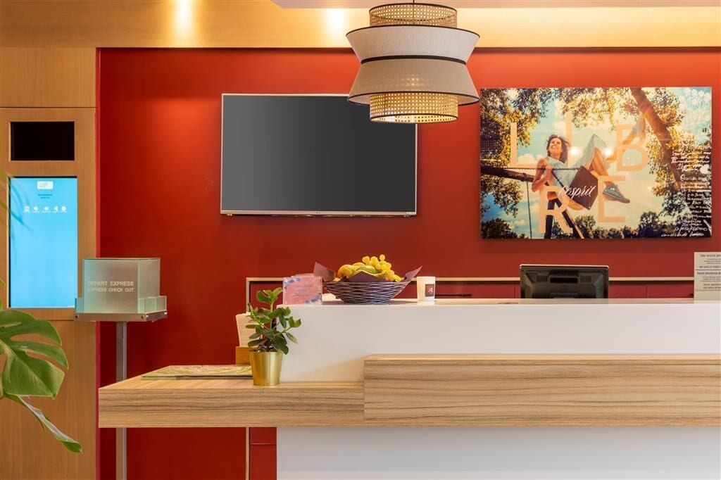Zona de recepció d'hotel amb una paret vermella, un televisor de pantalla plana, una caixa d'autoinformació digital, un taulell de recepció, una planta en test i un plat de fruita.