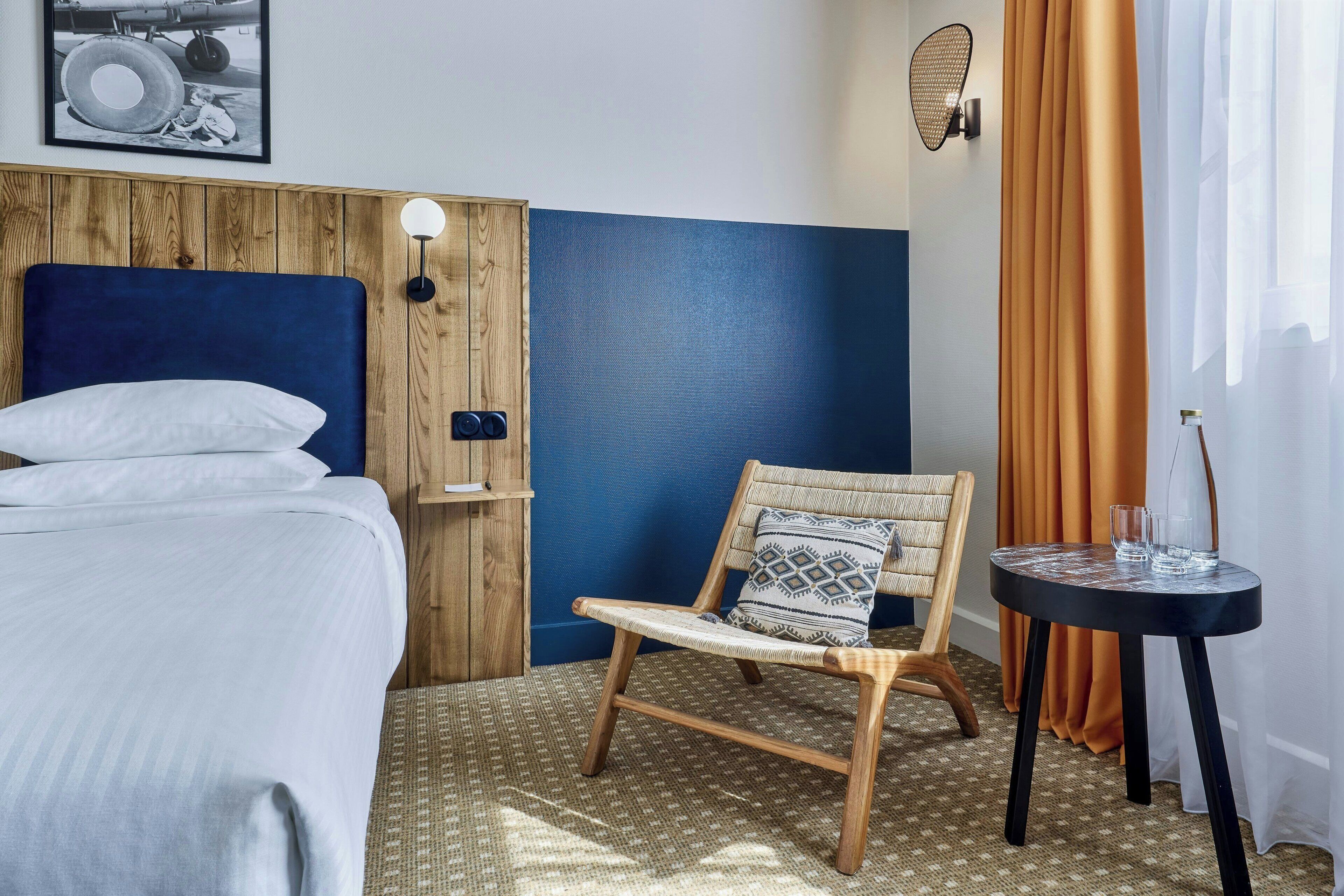 A hotel room with a bed, a chair, a small table, and a wall-mounted lamp.