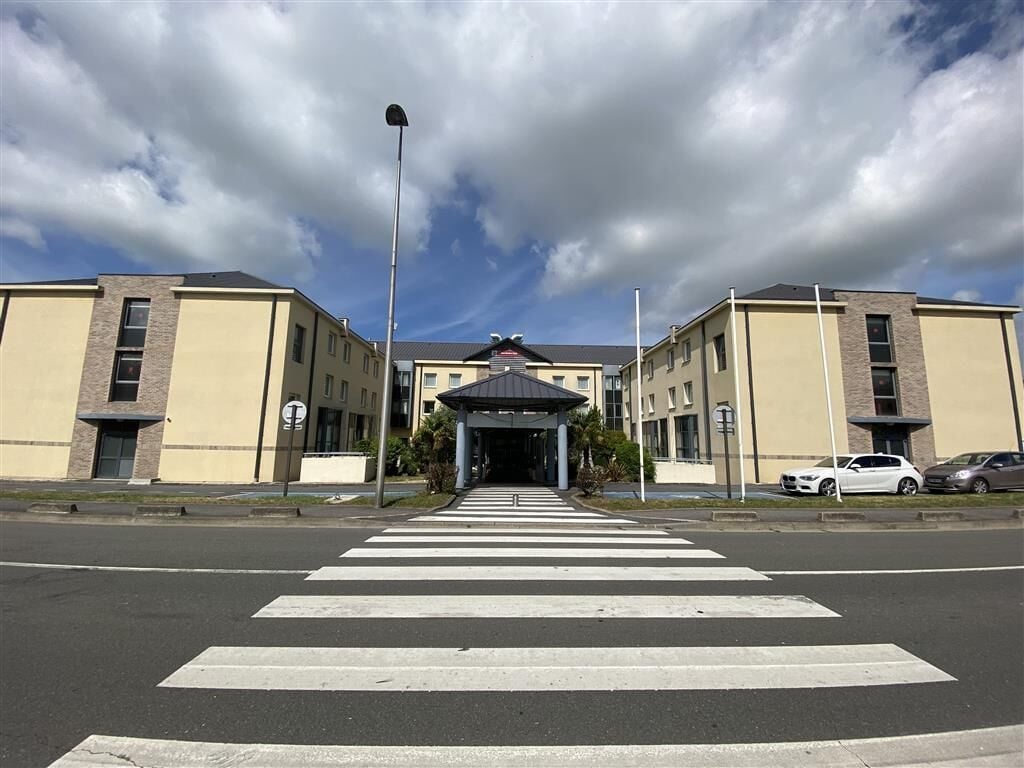 Een gebouw met een centrale ingang, een zebrapad ervoor en een parkeerplaats met auto's.