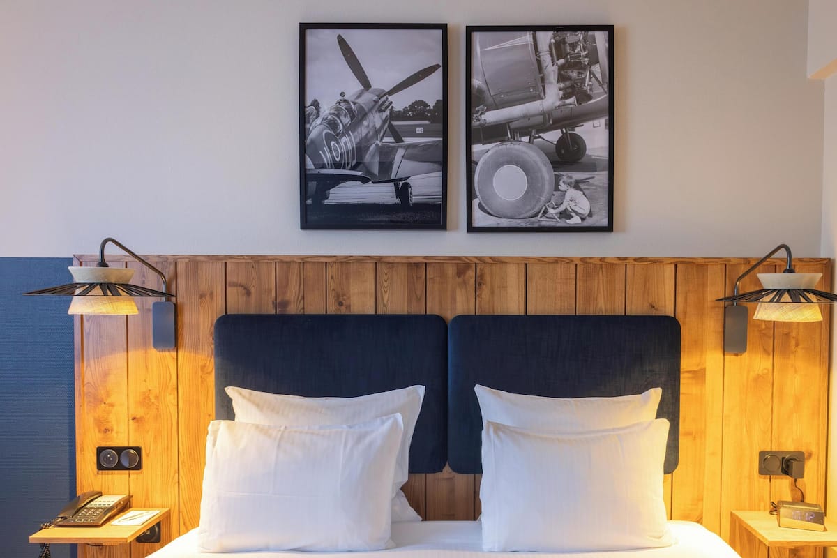 A hotel room with a wooden headboard, two bedside tables with lamps, a bed with white linens, and two framed pictures on the wall.