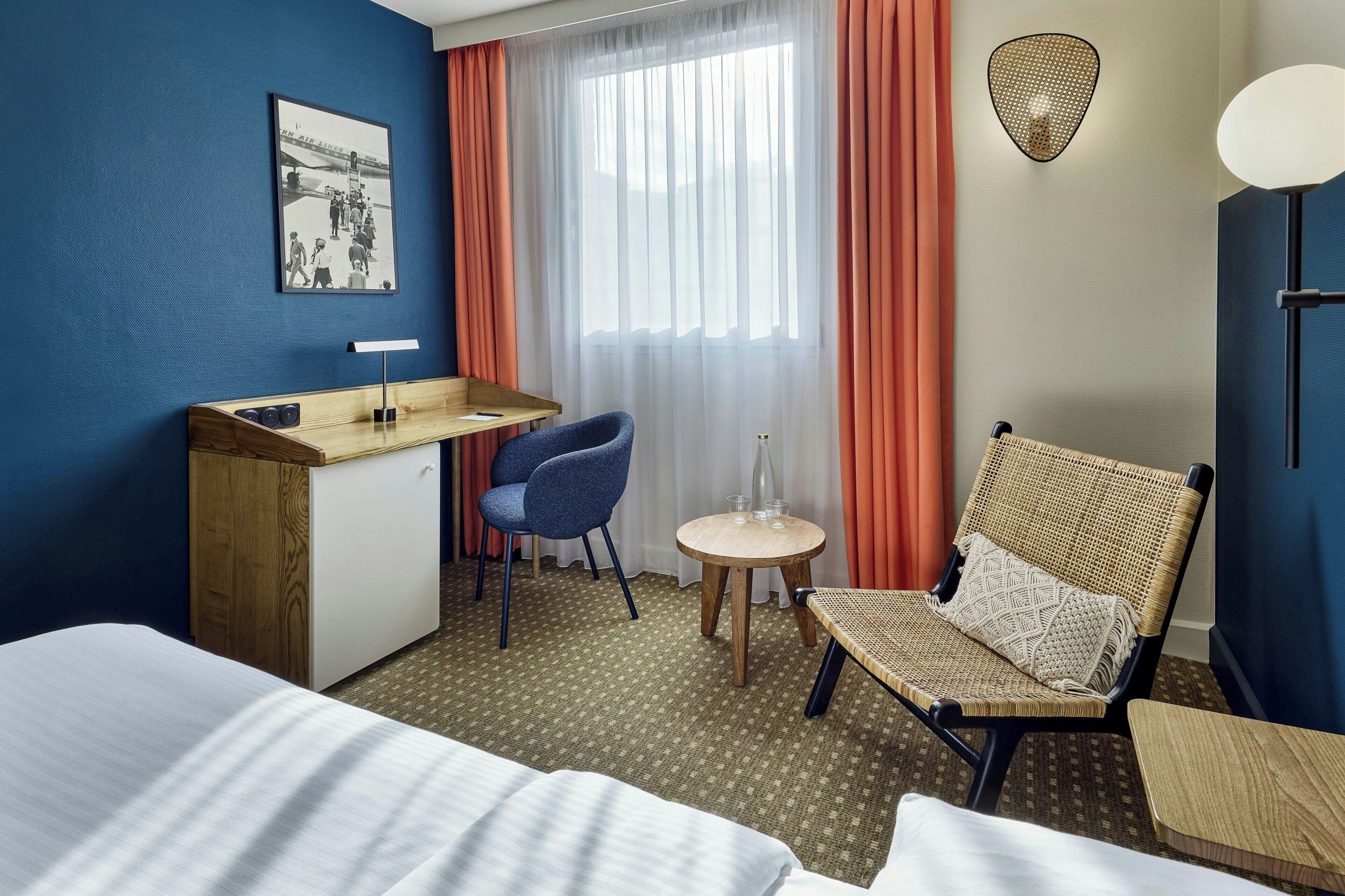 Habitación de hotel con cama, escritorio, silla, una mesita con vasos y una lámpara de pared.
