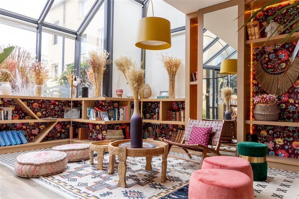 A room with a large window, a round table, and a bookshelf filled with books and decorative items.