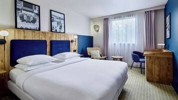 Habitación de hotel con una cama grande, un escritorio, una silla y un ventanal con cortinas.