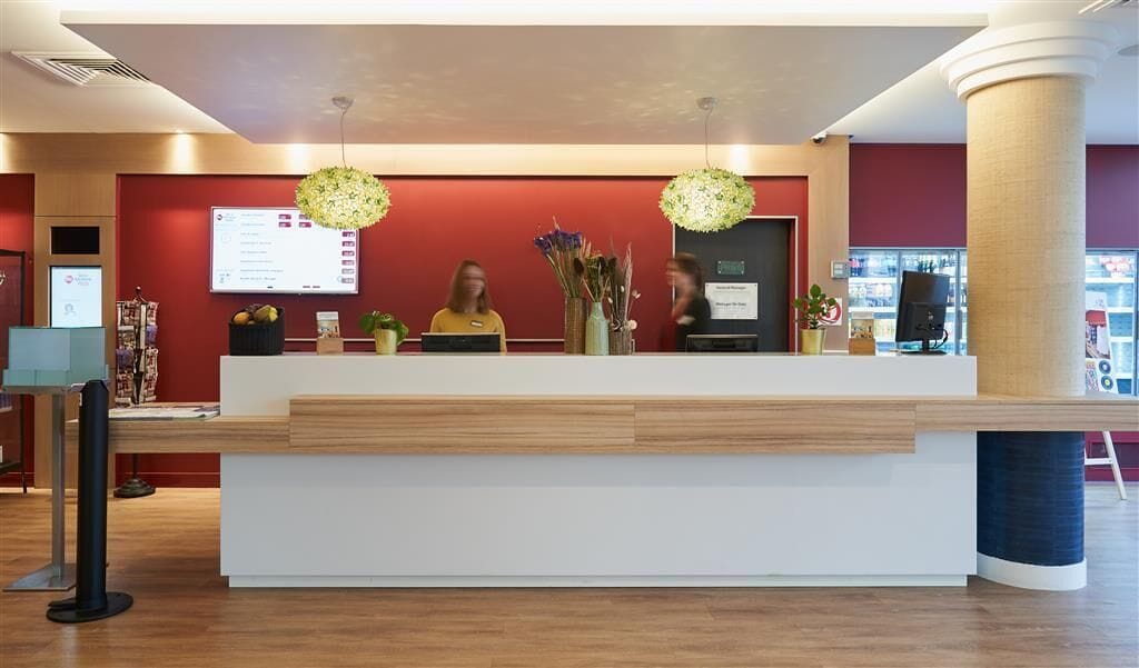 A reception desk with a digital display, two staff members, and a vase with flowers.