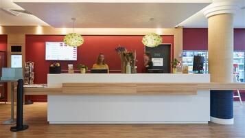 A reception desk with a digital display, two staff members, and a vase with flowers.