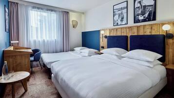 Une chambre d’hôtel avec deux lits, un bureau, une chaise et une petite table. On y trouve des rideaux, une lampe fixée au mur et deux tableaux encadrés accrochés au mur.