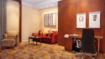 A hotel room with a red sofa, a wooden desk, a black office chair, a patterned carpet, and a large window.