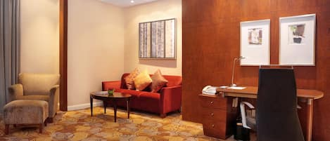 A hotel room with a red sofa, a wooden desk, a black office chair, a patterned carpet, and a large window.