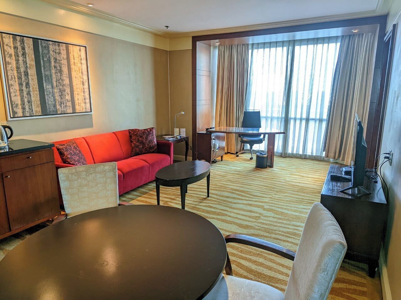 Habitación de hotel con sofá rojo, una mesa redonda, escritorio y un ventanal con cortinas.