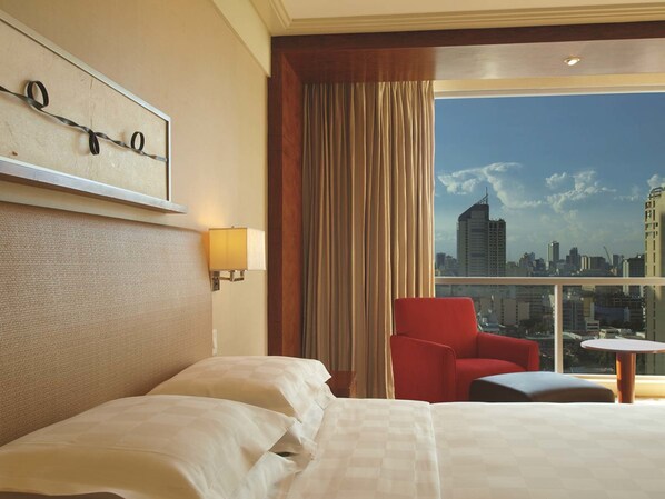 A hotel room with a large bed, a red armchair, a small round table, a city view through the window, and a wall-mounted lamp. - New Coast Hotel Manila (Manila)