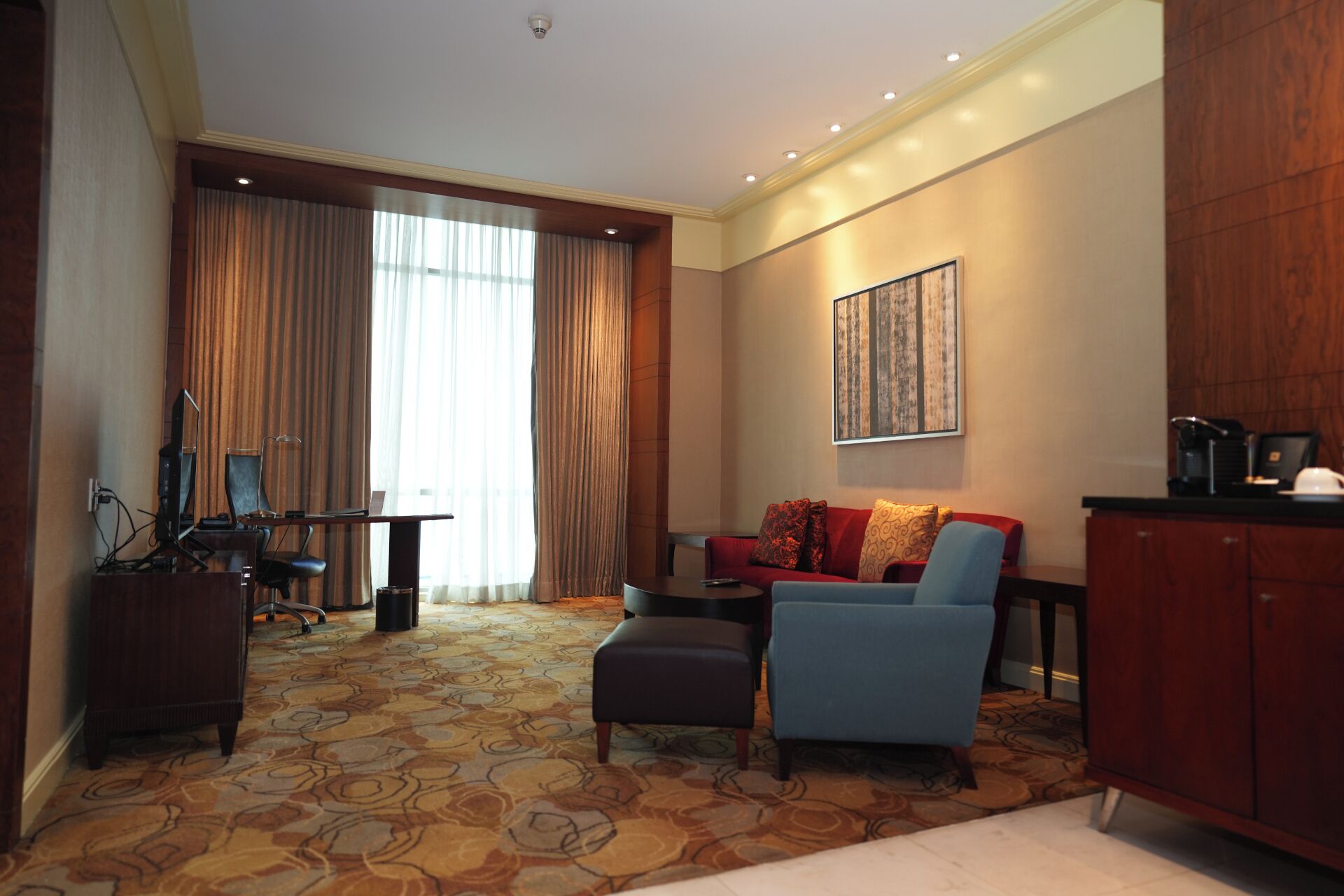A hotel room with a seating area, a desk with a computer, a large window with curtains, and a painting on the wall.