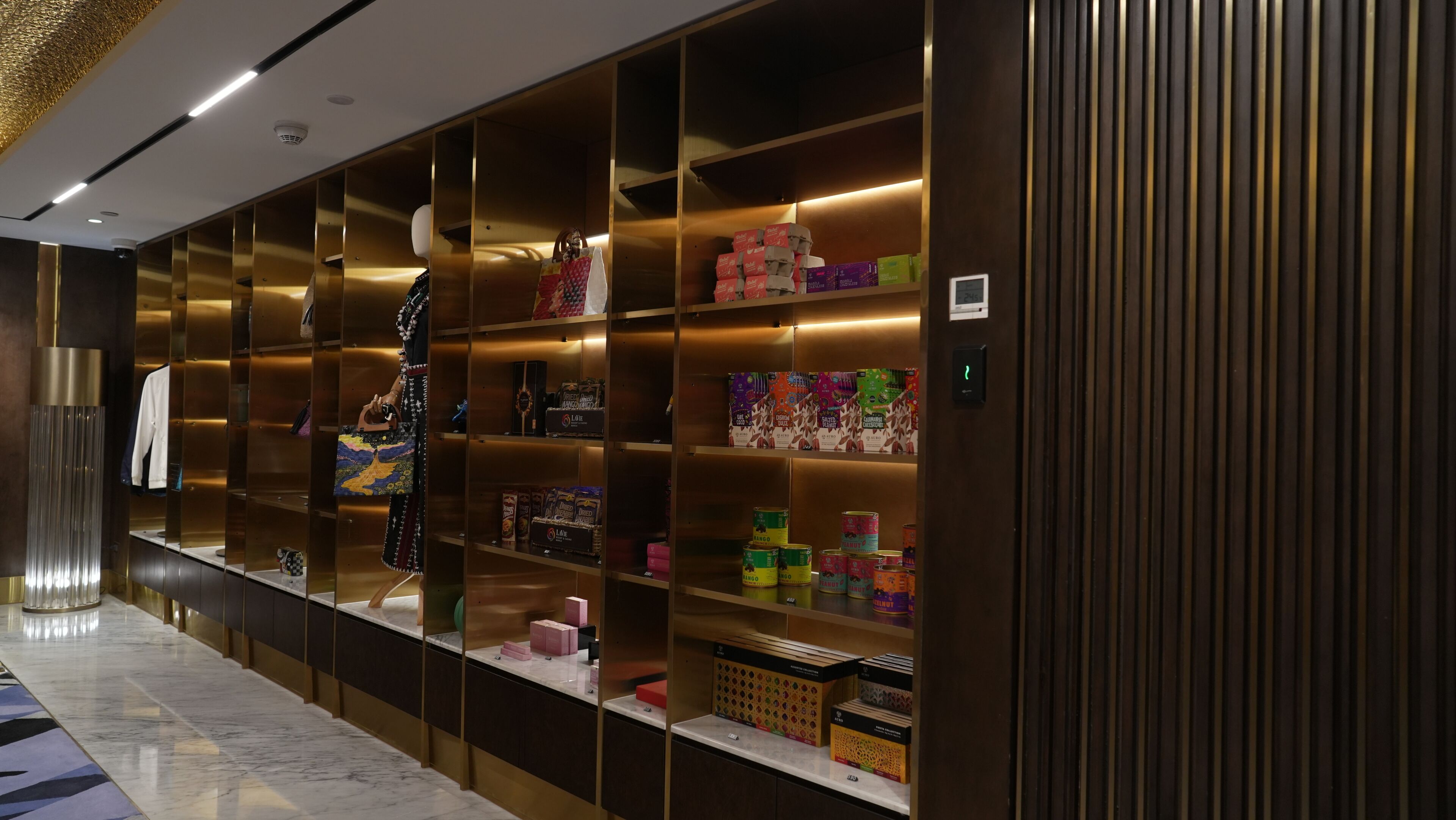 A hotel corridor with a series of shelves displaying various products, a mannequin dressed in a black outfit, and a marble floor.