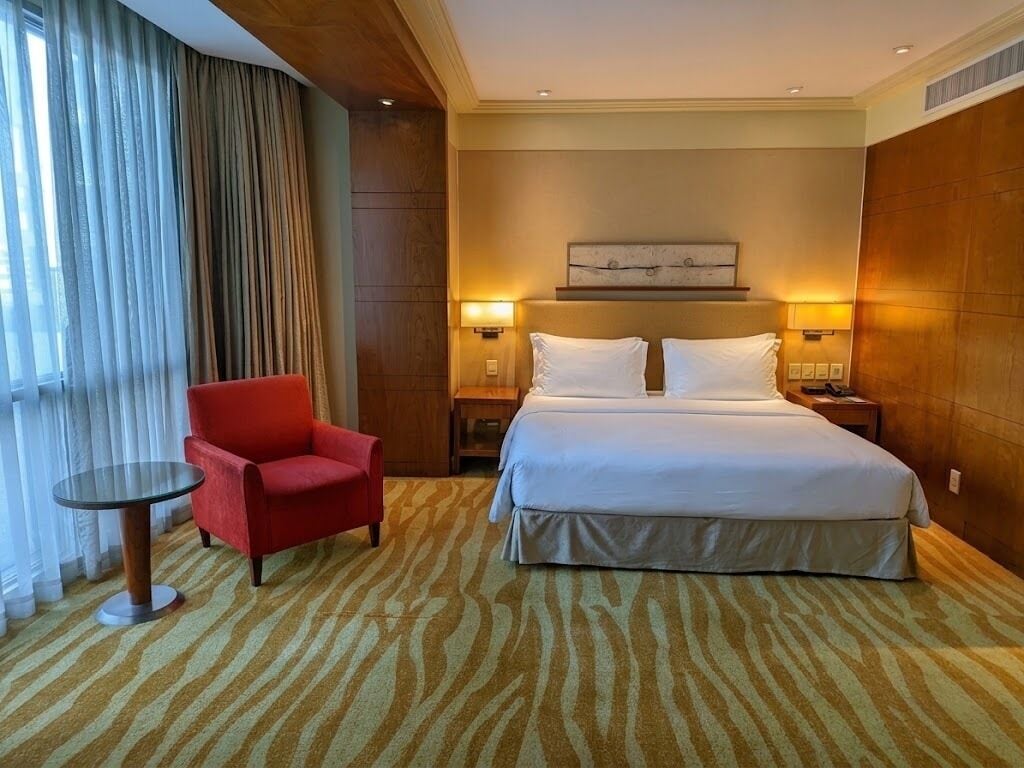 Habitació d'hotel amb un llit, un butac armchair vermell, una taula petita i dues làmpades de nit.