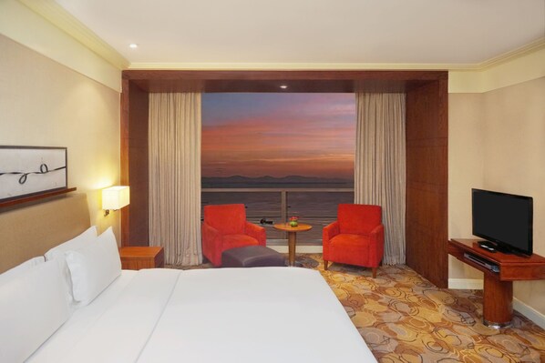A hotel room with a large bed, two red armchairs, a small table, a television on a wooden stand, and a view of the sunset through the window. - New Coast Hotel Manila (Manila)