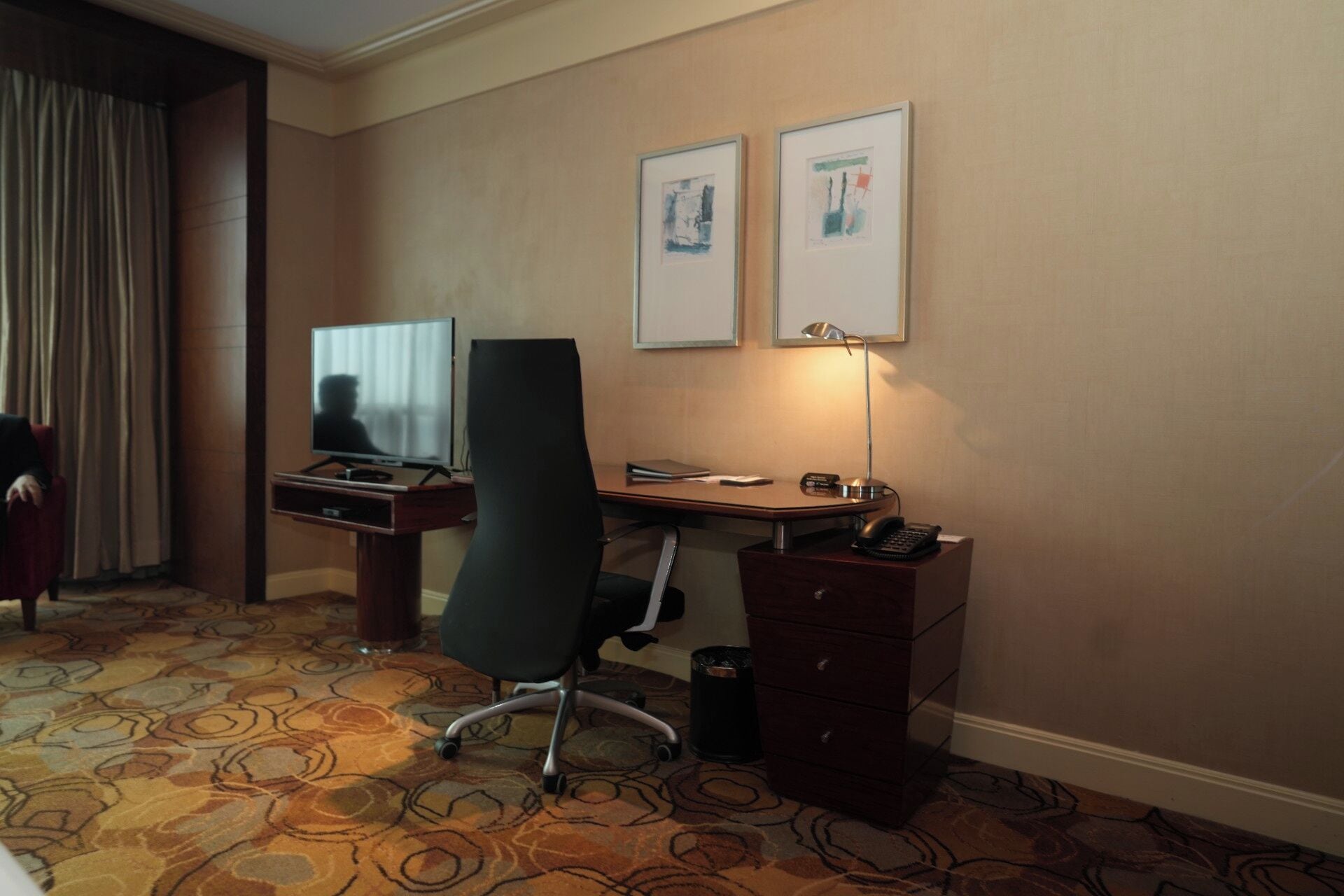 A hotel room with a desk, chair, television, lamp, and two framed pictures on the wall.