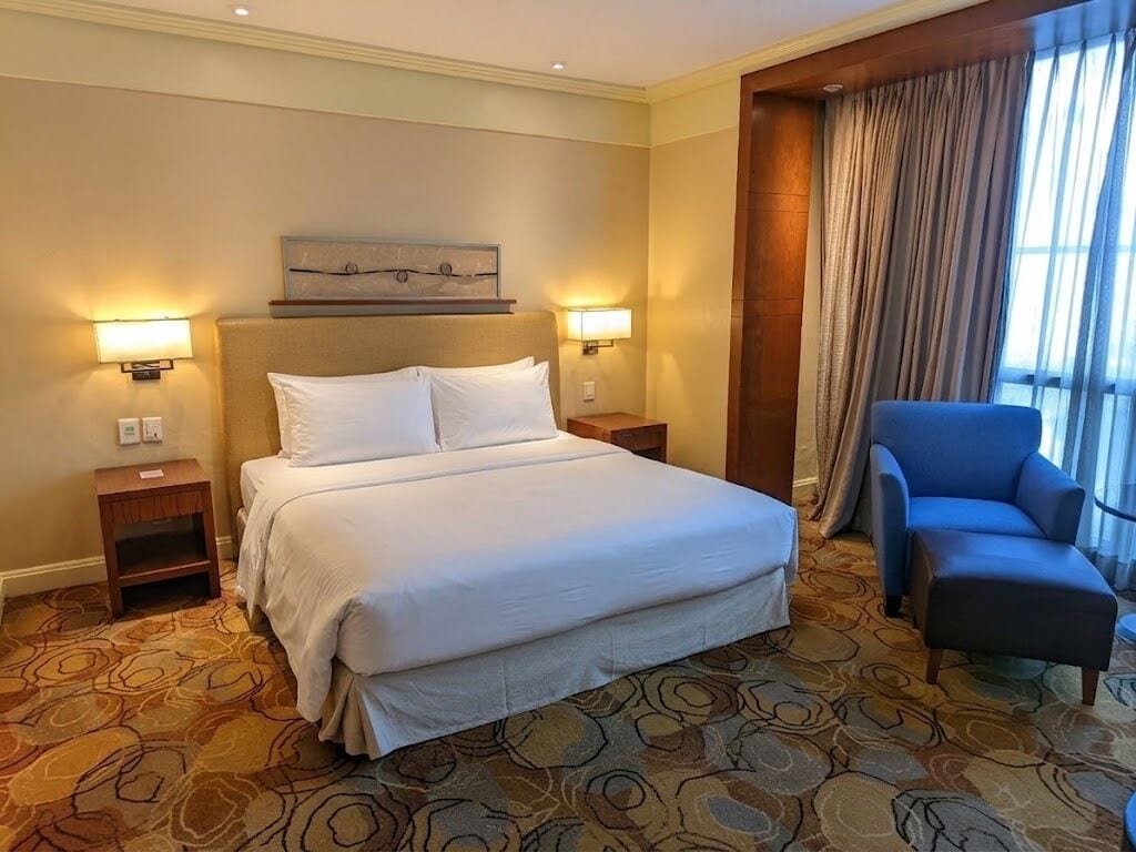 A hotel room with a large bed, two bedside lamps, a blue armchair, and a patterned carpet.