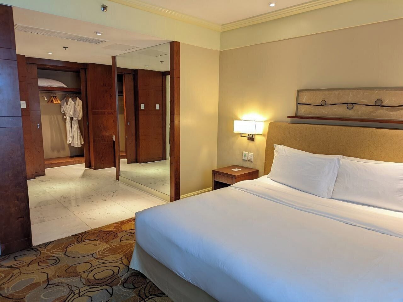 A hotel room with a large bed, a bedside table, a nightstand, a mirror, and a wardrobe.