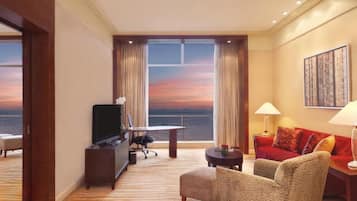 A hotel room with a large window offering a view of the ocean at sunset, a television, a desk, a sofa, and a lamp.